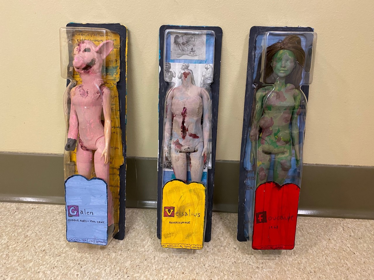 Three dolls depicting female bodies in unnatural conditions of mutilation or alteration. Each doll corresponds with a different medical authority and historical era.